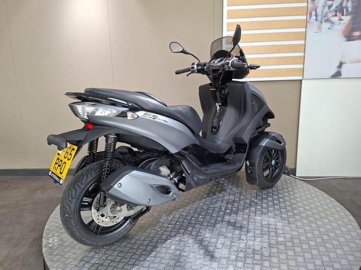 A black three-wheeled scooter displayed on a circular metallic platform, featuring a sleek design, a windshield, and a license plate. The background consists of a smooth, light-colored wall.