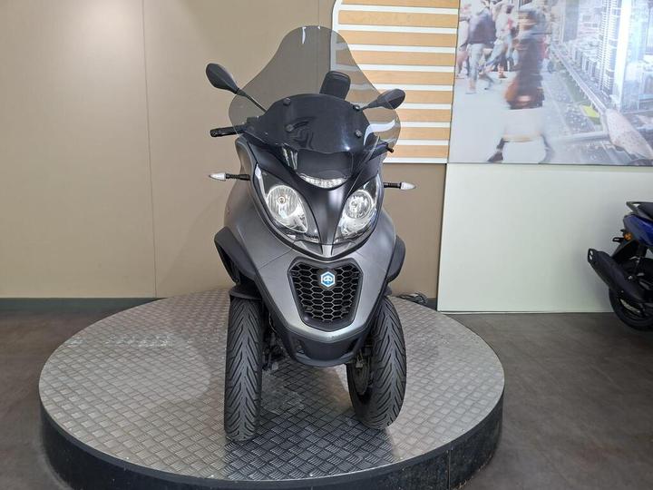 A gray three-wheeled scooter displayed on a circular platform, with a transparent windscreen and a modern design, against a neutral colored wall.