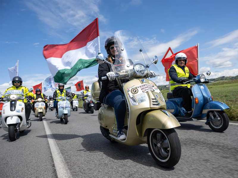 A group of scooter riders participating in a rally, with a woman in the foreground waving a Hungarian flag, while various flags fly from the scooters in a scenic outdoor setting.