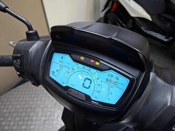 Close-up of a scooter's digital dashboard displaying speed, odometer reading of 6980 miles, and warning lights, against a blurred background of the scooter's body.