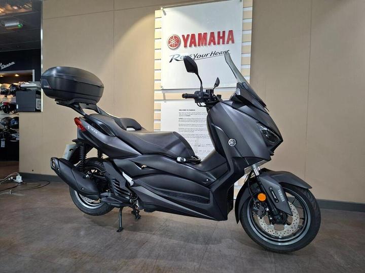 A sleek black Yamaha scooter displayed in a showroom, featuring storage on the back and a modern design. A promotional sign is visible in the background.