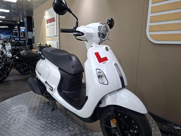 A white scooter with a learner's permit sticker parked on a display floor, surrounded by other motorcycles in a showroom.