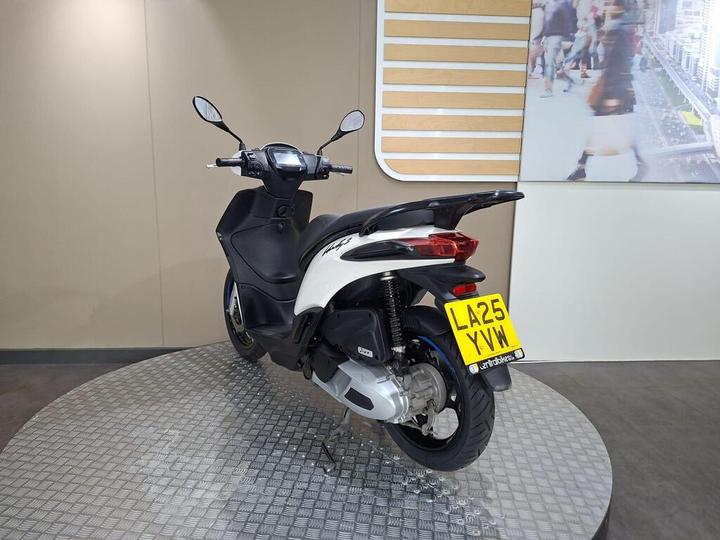 A white and black scooter displayed on a circular platform in a showroom, with a partial view of a wall featuring a digital screen showing people walking. The scooter's license plate reads "LA25 YVW."
