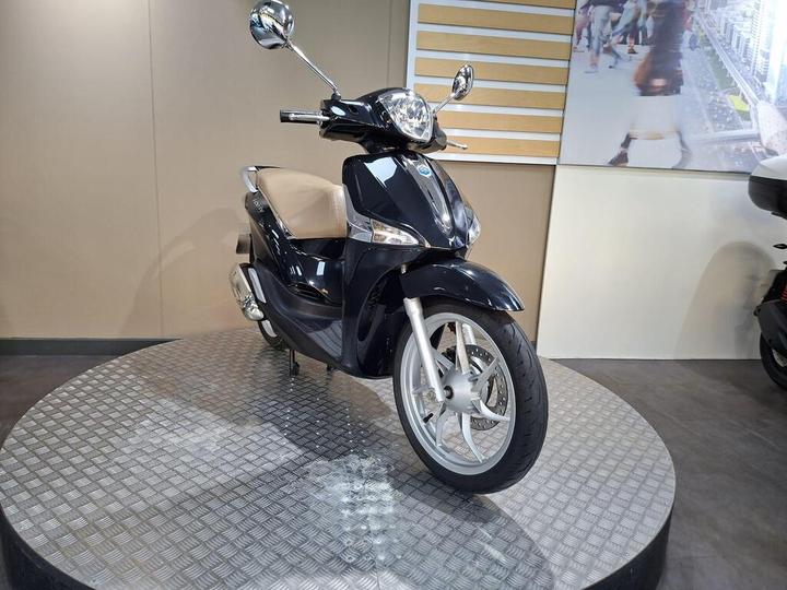 A black scooter with a beige seat is displayed on a rotating platform in a sleek showroom. The background features a modern wall with an image of a busy urban street.