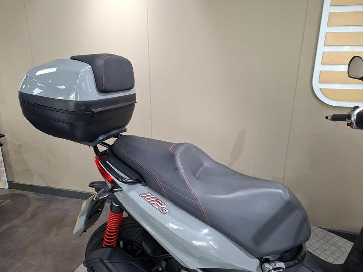 A side view of a motorcycle featuring a gray rear storage box and a sleek black seat with red stitching. The background is neutral and minimalistic, emphasizing the bike's design.