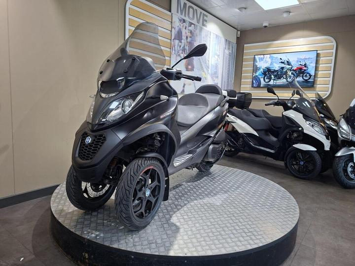 A black three-wheeled scooter displayed on a circular platform in a motorcycle showroom, with other scooters visible in the background.