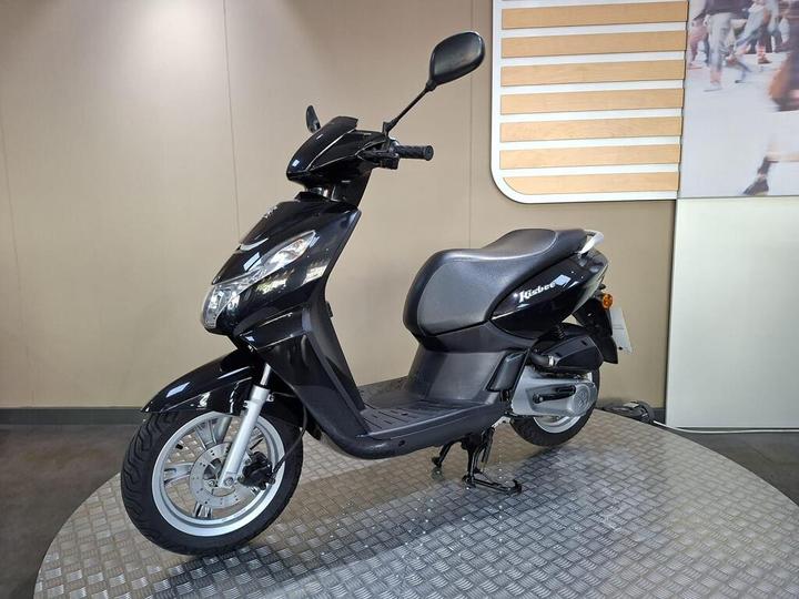 A black scooter is displayed on a stainless steel platform, with a minimalist background featuring wood paneling and a blurred image of people walking.