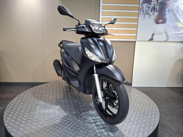 A sleek black scooter displayed on a round, textured platform against a neutral background.