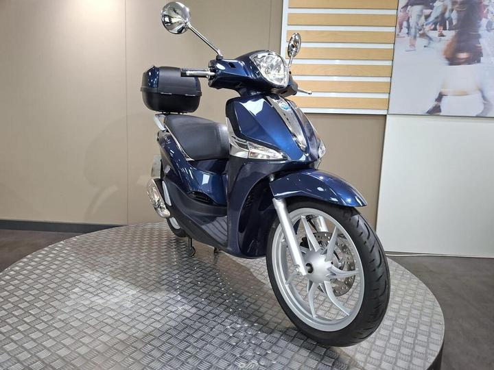 A blue scooter displayed on a round platform with a textured surface, featuring a rounded front, a single rearview mirror, and a black storage box attached to the back. The background includes a light-colored wall and a large image of people walking.