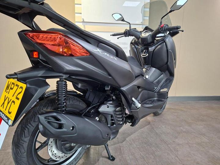 A close-up view of a black Yamaha XMAX scooter, highlighting the rear, exhaust, and rear suspension. The image includes a license plate with the number "MP72 XYX" and an indoor showroom setting with a reflection in the background.