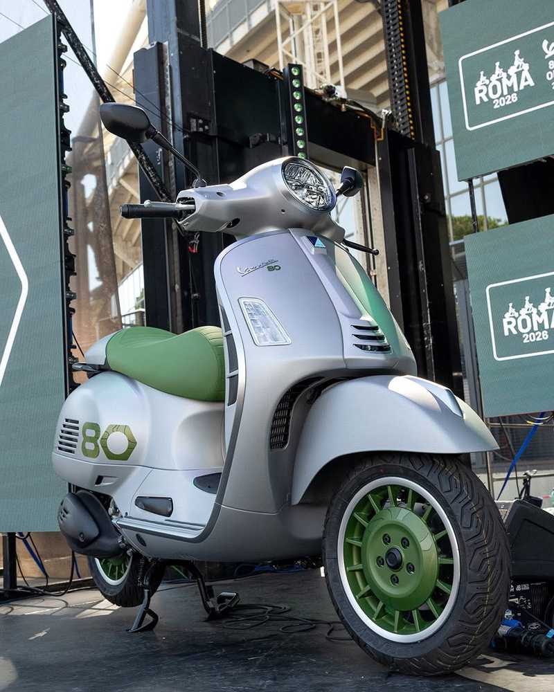 A silver and green Vespa scooter is displayed prominently on stage, with digital screens in the background promoting an event for Roma 2026.