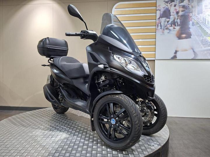 A black three-wheeled scooter displayed on a textured platform, featuring a large windshield, comfortable seat, and a rear storage compartment. Background includes a blurred image of people walking.