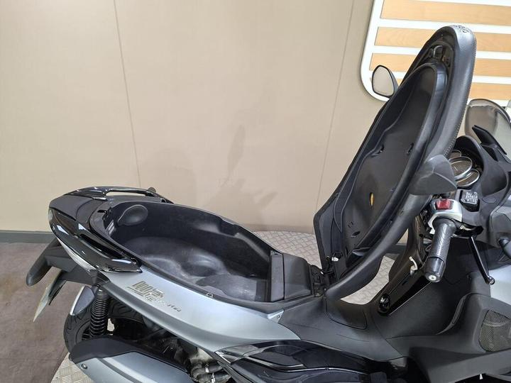 A close-up view of a scooter with its seat raised, revealing the storage compartment beneath. The scooter features a sleek silver and black design, showcasing the handlebars and part of the tire in the foreground. The background is plain and neutral-colored.
