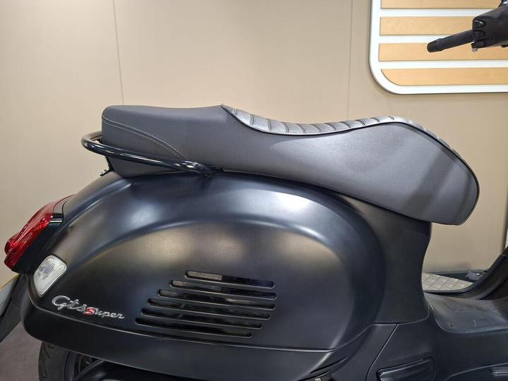 A close-up of the rear section of a black scooter, showcasing the matte finish, sleek design, and textured seat with stitched detailing. The scooter emblem "GTS Super" is visible on the side. The background is neutral and unobtrusive.