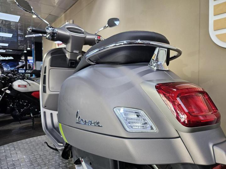 A close-up view of a grey Vespa scooter showcasing its rear design, taillight, and chrome accents, with a blurred background of other motorcycles in a showroom setting.