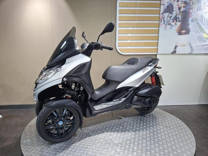 A silver and black three-wheeled scooter displayed on a circular platform against a neutral background, featuring a transparent windscreen and a sleek design.