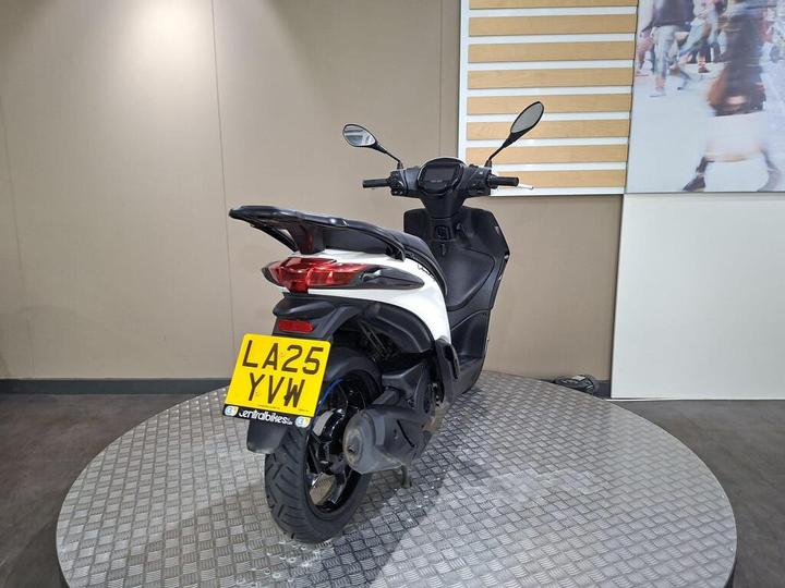 A black and white scooter is displayed on a circular checkered platform, showing its rear side and license plate reading "LA25 YVW". The background features a plain wall and a partially visible wall image depicting people.