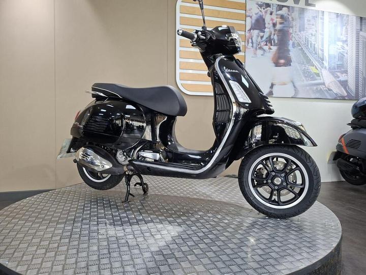 A black Vespa scooter displayed on a circular metallic platform, with a minimalist backdrop featuring urban imagery.