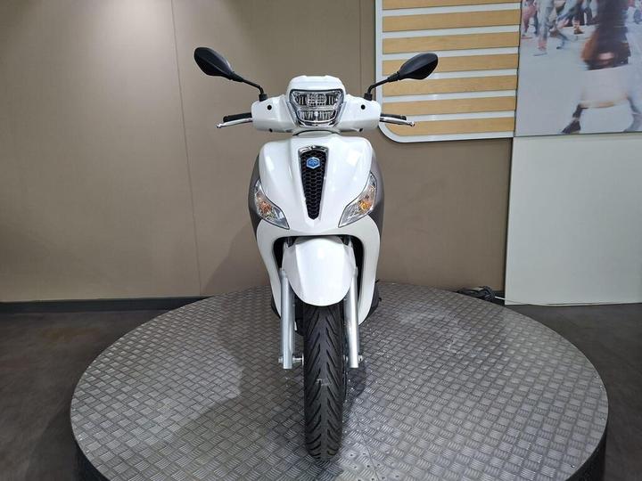 A white scooter displayed frontally on a circular showroom platform, with a simple background featuring a light-colored wall and a blurred image of people in motion.