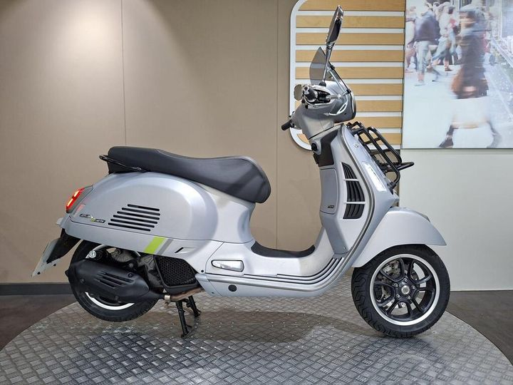 Vespa GTS 125 Supertech (2023) A silver scooter displayed on a circular platform against a neutral backdrop, featuring a sleek design and modern details.