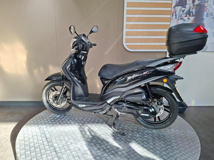 A black SYM scooter displayed on a circular platform with a textured surface, featuring a sleek design and a rear storage box. The background is minimal and softly lit.