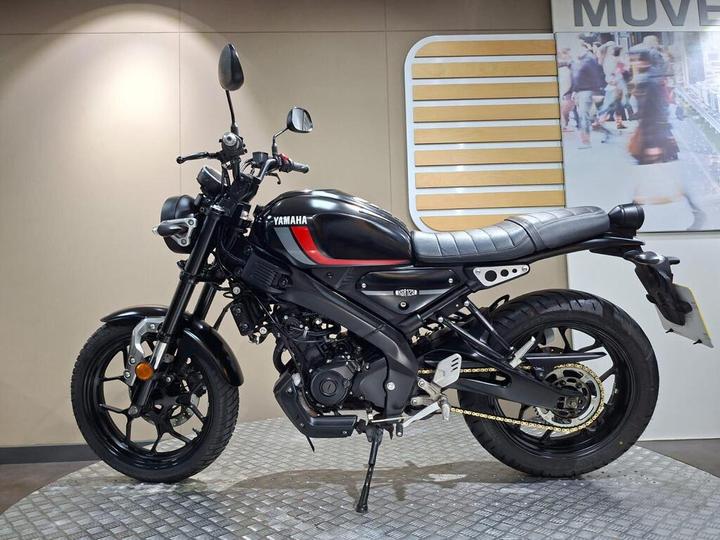 A Yamaha motorcycle displayed on a diamond-patterned platform, featuring a black frame with red accents, a black seat, and sleek design elements, against a neutral background with minimal lighting.