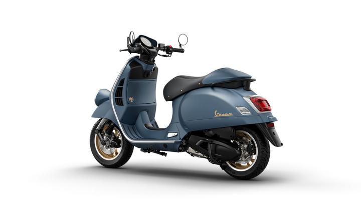 A blue Vespa scooter with a sleek design and gold accents, viewed from the rear-right angle, showcasing its modern aesthetic and stylish curves.