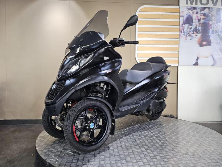 A black three-wheeled scooter displayed on a circular platform, featuring a large windshield and sporty wheels with red accents. The background includes a subtle patterned wall and a blurred image of people walking.
