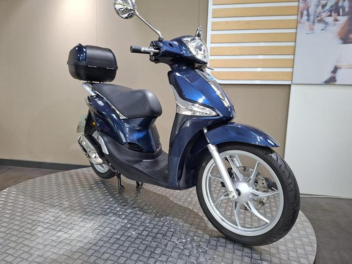 A blue scooter with a black storage box on the rear, positioned on a textured surface, against a neutral background.