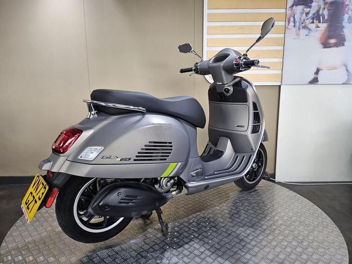 A sleek gray scooter displayed on a round metallic platform, showcasing its rear and side profile, with a modern design and subtle green accents. The background features a soft-focus image of people walking.