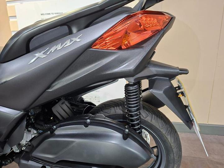 Close-up of the rear section of a Yamaha XMAX scooter, showing the rear suspension, taillight, and XMAX logo against a light-colored background.