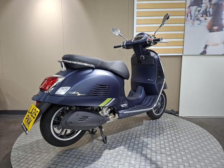 A blue Vespa scooter is displayed on a circular metal platform with a textured surface, showcasing its sleek design and modern features. The background is a neutral tan color with a minimalist design.