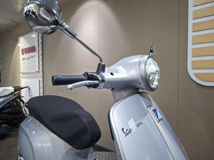 A close-up of a silver Vespa scooter with a round headlight, mirror, and handlebar against a neutral-colored wall.