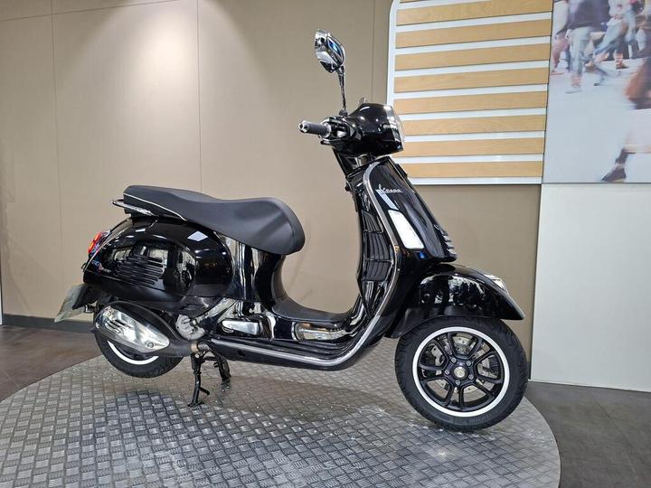Black scooter with sleek design displayed on a circular patterned floor against a neutral background.