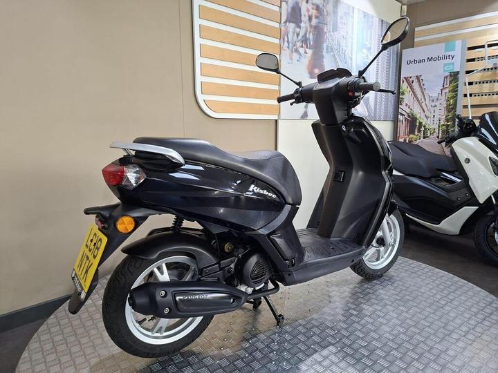 A black scooter with a sleek design, featuring a small windscreen, side mirrors, and a prominent rear light. It is showcased on a metal floor in an indoor setting, with a blurred background displaying urban scenery and another scooter.