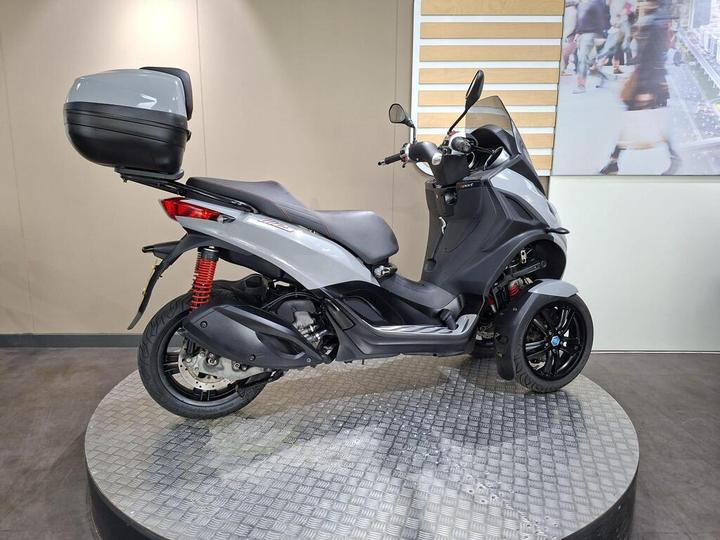 A sleek gray three-wheeled scooter displayed on a round platform, featuring a rear storage box and a modern design. The background includes a light-colored wall with a visual element depicting a city scene.