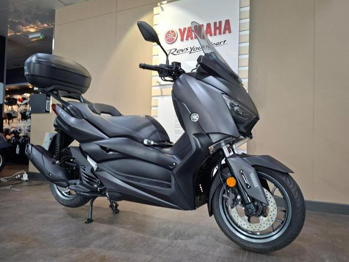 A sleek, matte black Yamaha scooter displayed in a showroom, featuring a large rear storage compartment and prominent branding in the background.