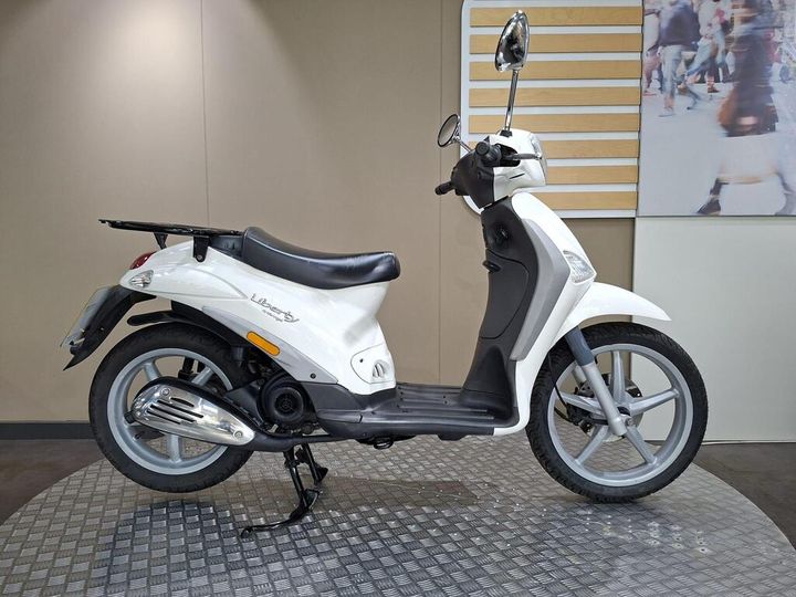 Piaggio Liberty 50 Delivery (2015) A white scooter is positioned on a patterned surface, displaying its side profile. The scooter features a black seat, chrome exhaust, and a rear luggage rack, with a neutral background that includes light-colored walls.
