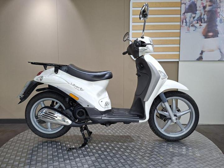 Piaggio Liberty 50 Delivery (2015) A white scooter is positioned on a patterned surface, displaying its side profile. The scooter features a black seat, chrome exhaust, and a rear luggage rack, with a neutral background that includes light-colored walls.