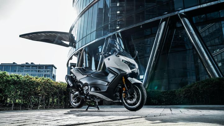A sleek, modern gray motorcycle is prominently displayed in front of a contemporary glass building, with sunlight illuminating its design and reflections visible on the building's surface.