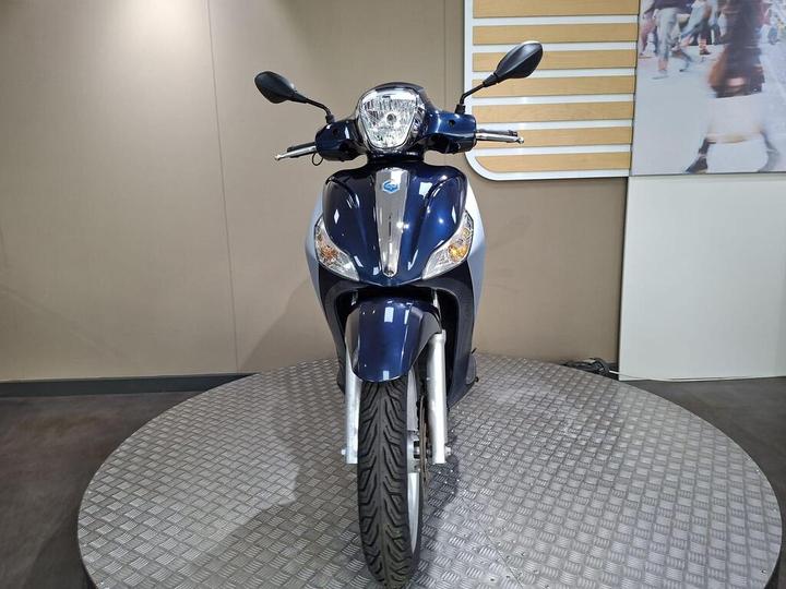 A front view of a blue scooter displayed on a circular metallic platform against a plain background. The scooter features a shiny headlight and sleek design, emphasizing its front profile.