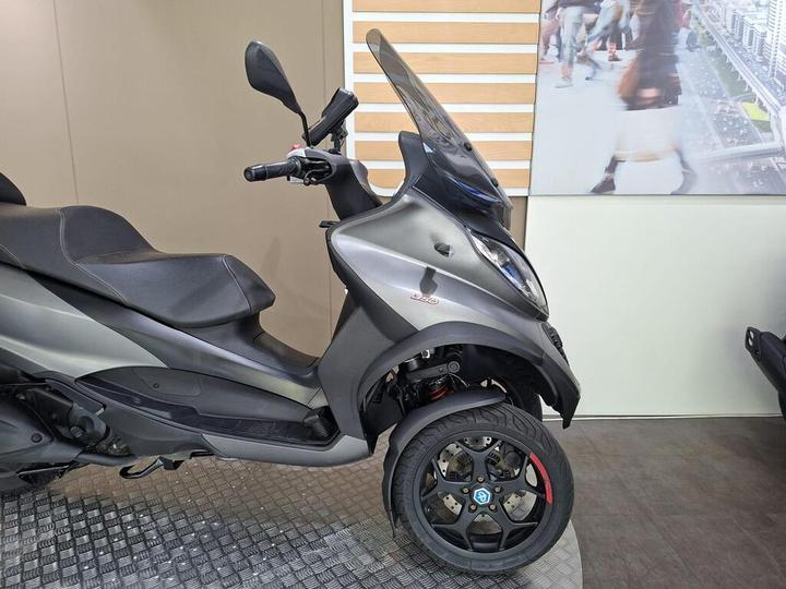 A parked gray scooter with a sleek design, featuring two front wheels and a black seat, positioned on a textured floor in a showroom.