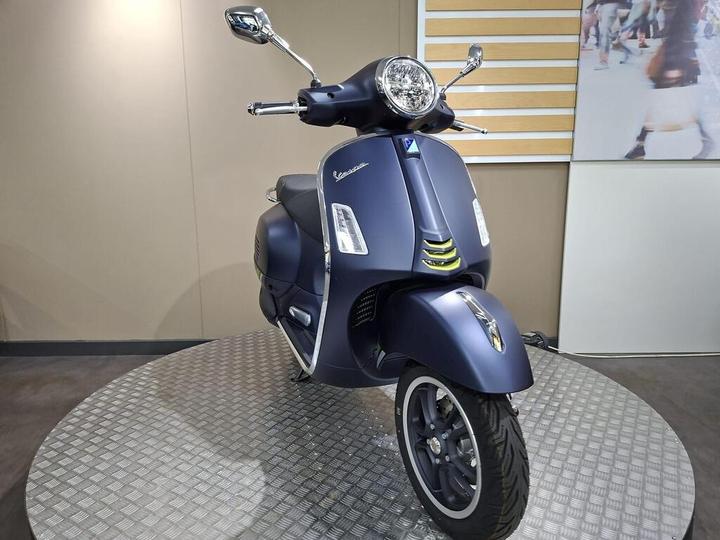 A dark blue Vespa scooter displayed on a rotating platform with a modern background, showcasing its sleek design and chrome accents.