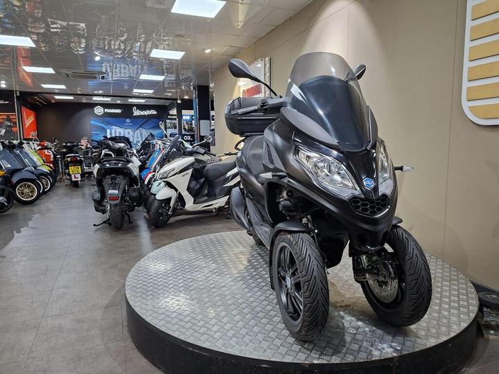 A black three-wheeled scooter is displayed on a circular platform in a modern showroom, surrounded by various other scooters and motorcycles. The background features branding for Piaggio and Vespa, along with a visible salesperson in the corner.