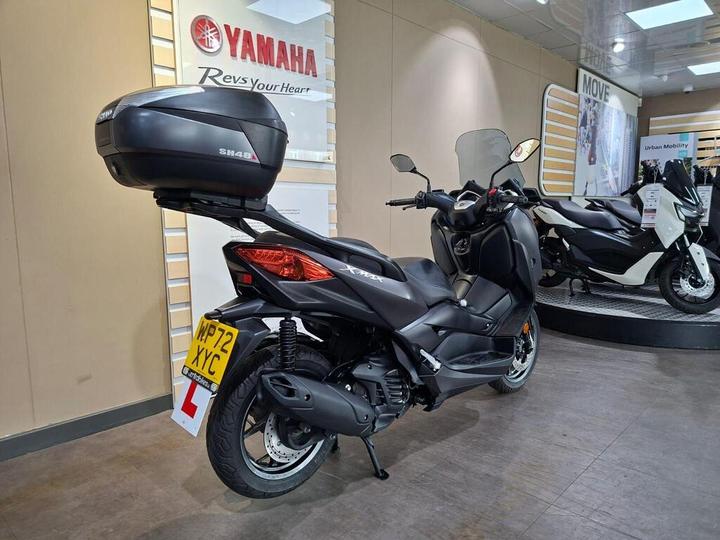 A black Yamaha scooter parked indoors, featuring a rear top box, with a visible learner plate and a display of other scooters in the background.