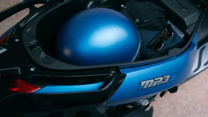 A blue motorcycle helmet with a hexagonal pattern is placed inside the storage compartment of a motorcycle, alongside a pair of sunglasses, black gloves, and a folded piece of clothing. The motorcycle features a stylish blue and black design with the model name "MP3" visible.