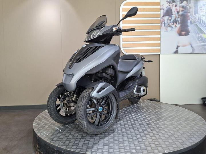 A modern gray three-wheeled scooter displayed on a round platform with a textured surface, set against a neutral background featuring a blurred image of busy pedestrians.