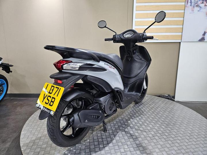 A silver scooter with a distinctive design parked on a textured circular platform, showcasing its rear and side profile. The license plate is visible, and the background features a neutral wall.