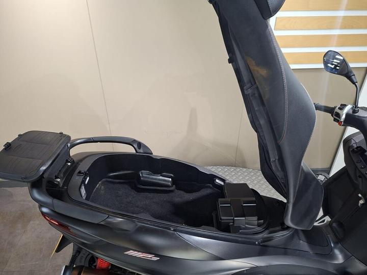 A close-up view of the storage compartment of a scooter, featuring an opened seat that reveals the interior, with a dark carpeted floor and a small storage unit on the left side.