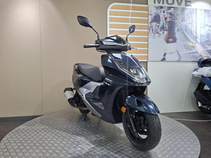 A dark blue modern scooter displayed on a circular platform in a well-lit showroom, with a background featuring a large image of people and urban scenery.